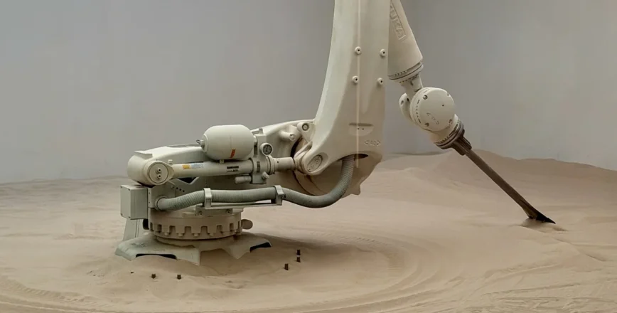 Un bras robotique de couleur claire est posé sur un terrain sablonneux dans un environnement intérieur.