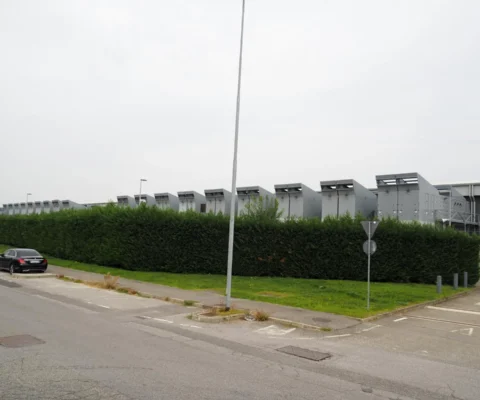 Un bâtiment industriel moderne avec une haie verte devant, sous un ciel gris.