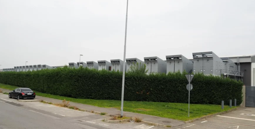 Un bâtiment industriel moderne avec une haie verte devant, sous un ciel gris.